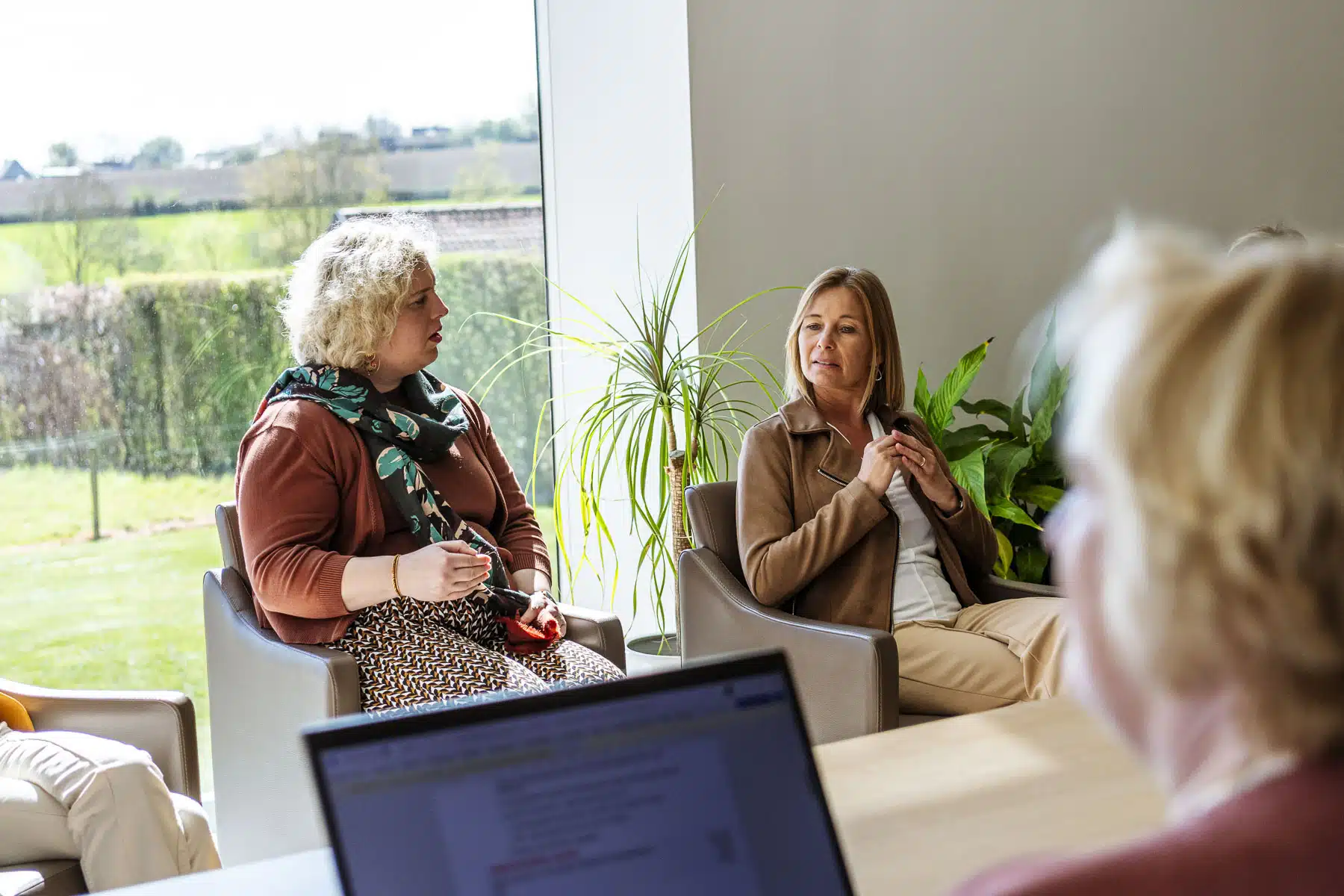 Twee vrouwen in gesprek tijdens juridische consultatie bij notariskantoor, zittend bij raam met groene achtertuin
