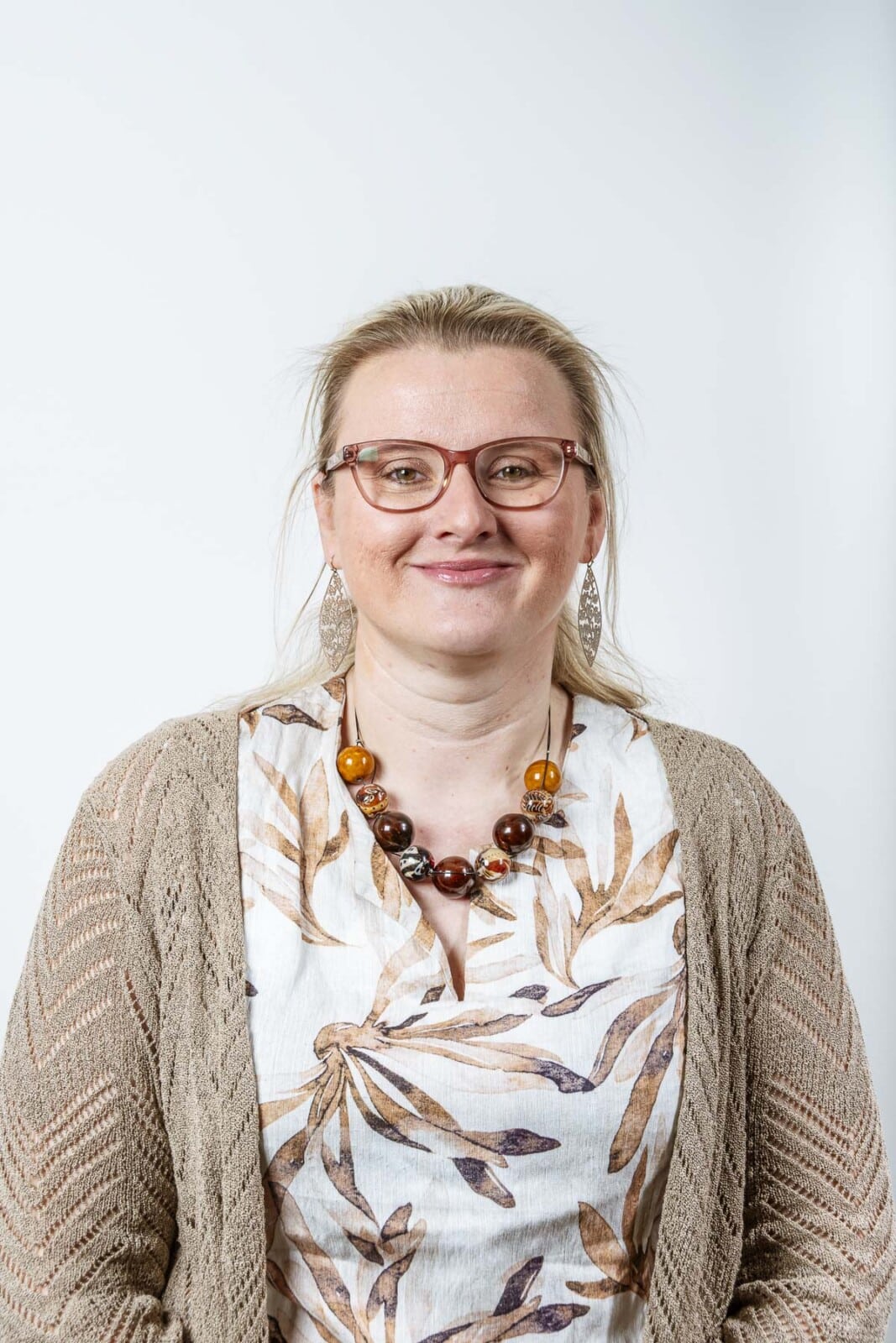 Professionele vrouw met blonde haar, bril en beige vest glimlacht vriendelijk naar de camera tegen witte achtergrond