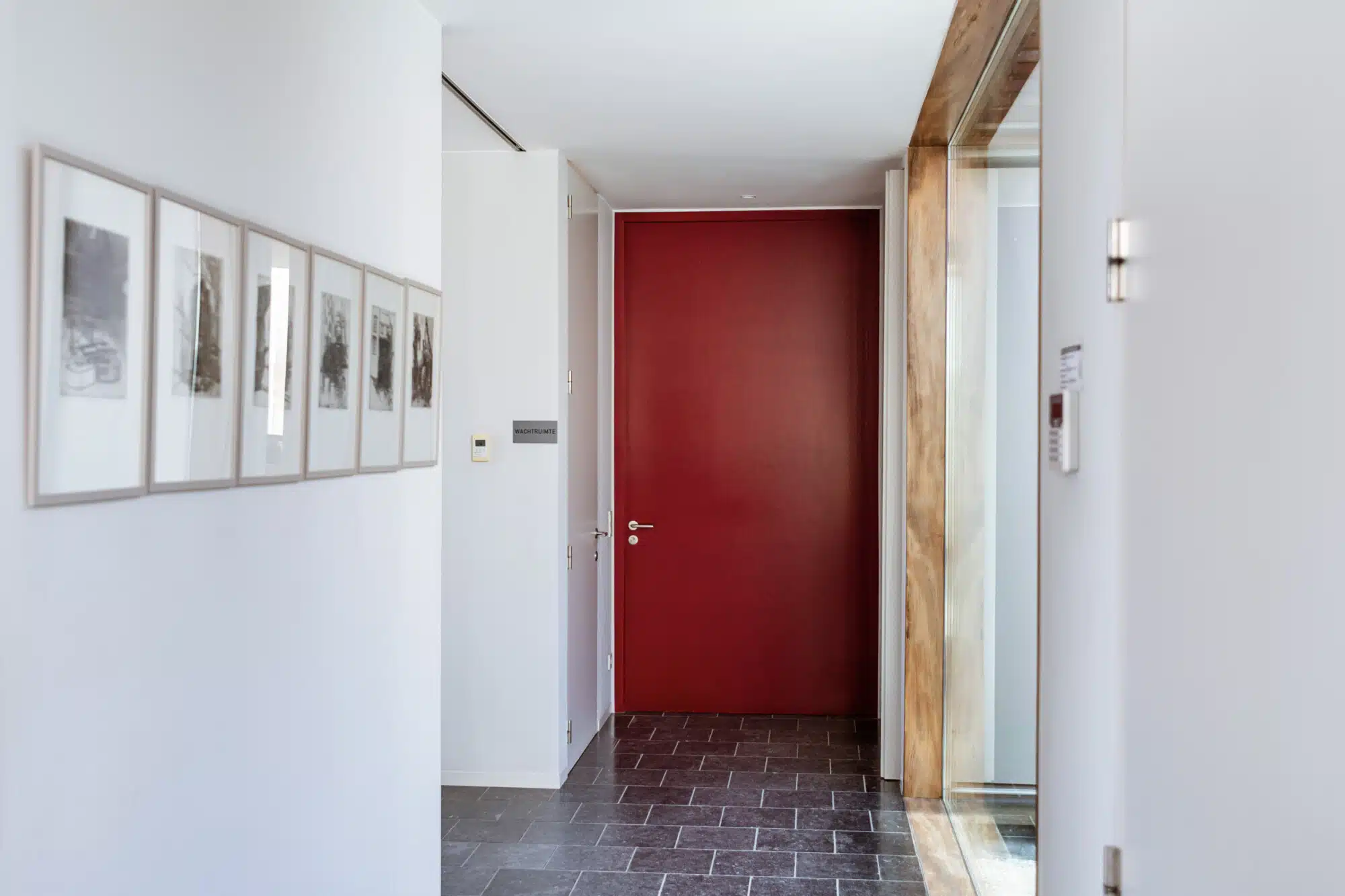 Moderne kantoorgang met rode deur, houten balken en ingelijste kunstwerken aan witte muren